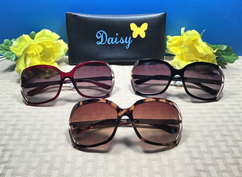 Op de afbeelding: Drie paar oversized zonnebrillen met verschillende kleuren frames. De frames zijn rood, zwart en schildpad. De zonnebrillen liggen op een wit oppervlak met een zwarte etui erachter. De etui heeft het woord "Daisy" en een vlinderontwerp erop.