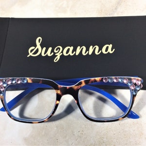 May include: Eyeglasses with a tortoiseshell pattern frame, blue arms, and embellished with pink and blue rhinestones. The glasses are paired with a black case with the name "Suzanna" in gold script.