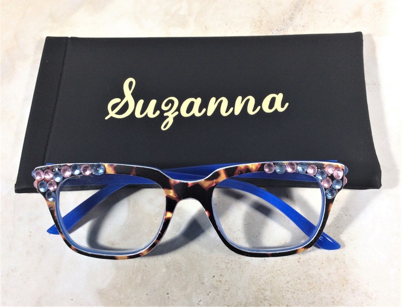 May include: Eyeglasses with a tortoiseshell pattern frame, blue arms, and embellished with pink and blue rhinestones. The glasses are paired with a black case with the name "Suzanna" in gold script.
