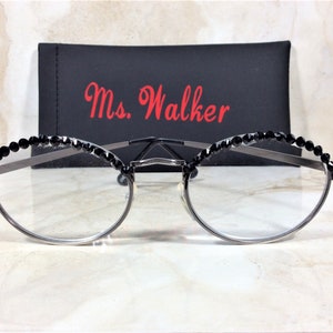 May include: Silver-framed eyeglasses with clear lenses, adorned with black crystal embellishments along the top rim. A black case with the name "Ms. Walker" in red script is behind the glasses. The glasses have a modern, elegant design.