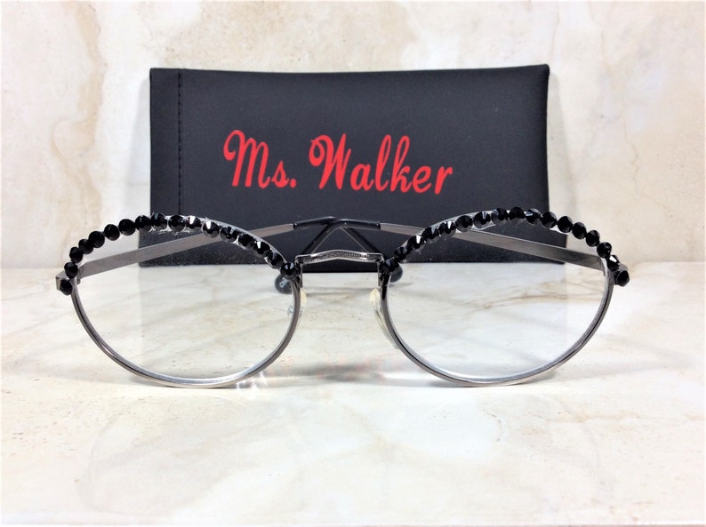May include: Silver-framed eyeglasses with clear lenses, adorned with black crystal embellishments along the top rim. A black case with the name "Ms. Walker" in red script is behind the glasses. The glasses have a modern, elegant design.