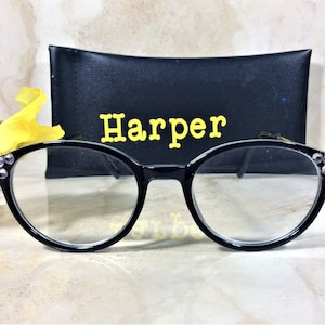 Puede incluir: Gafas con montura negra y lentes transparentes, adornadas con pedrer&iacute;a en las patillas. Se encuentran frente a un estuche negro con el nombre "Harper" en letras amarillas. El fondo incluye flores.