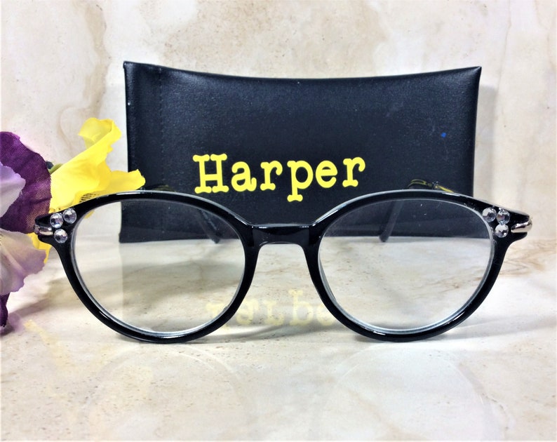 Puede incluir: Gafas con montura negra y lentes transparentes, adornadas con pedrer&iacute;a en las patillas. Se encuentran frente a un estuche negro con el nombre "Harper" en letras amarillas. El fondo incluye flores.
