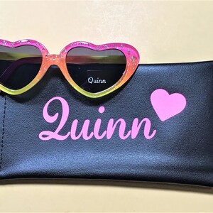 May include: Pink heart-shaped sunglasses with a rainbow gradient and glitter accents. The sunglasses are in a black case with the name "Quinn" and a pink heart printed on it.