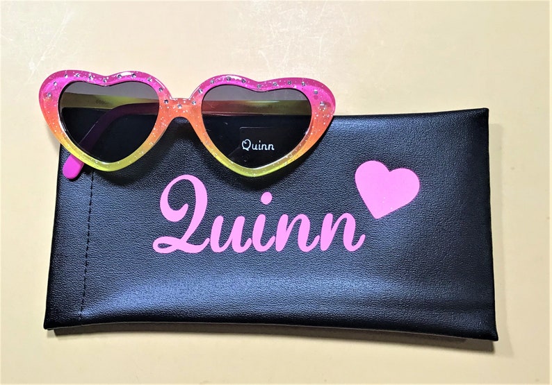 May include: Pink heart-shaped sunglasses with a rainbow gradient and glitter accents. The sunglasses are in a black case with the name "Quinn" and a pink heart printed on it.