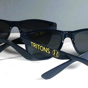 Puede incluir: Dos pares de gafas de sol azul marino oscuro con lentes oscuras. Las patillas llevan el texto amarillo TRITONS con un gr&aacute;fico de b&eacute;isbol. Un par tiene el nombre HUNTER #16 impreso en amarillo, mientras que el otro muestra JOSEPH #9 en el interior.