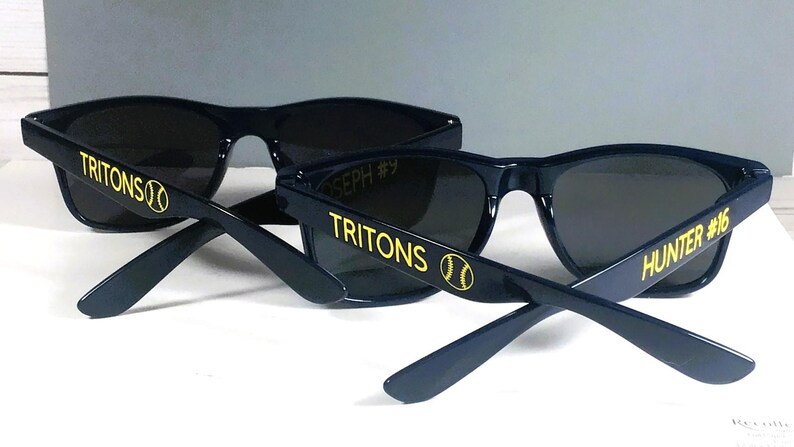 Puede incluir: Dos pares de gafas de sol azul marino oscuro con lentes oscuras. Las patillas llevan el texto amarillo TRITONS con un gr&aacute;fico de b&eacute;isbol. Un par tiene el nombre HUNTER #16 impreso en amarillo, mientras que el otro muestra JOSEPH #9 en el interior.