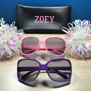 May include: Two pairs of sunglasses, one pink and one purple, with a black case that says "ZOEY" on it. The sunglasses are on a wooden surface with white and iridescent bows on either side.