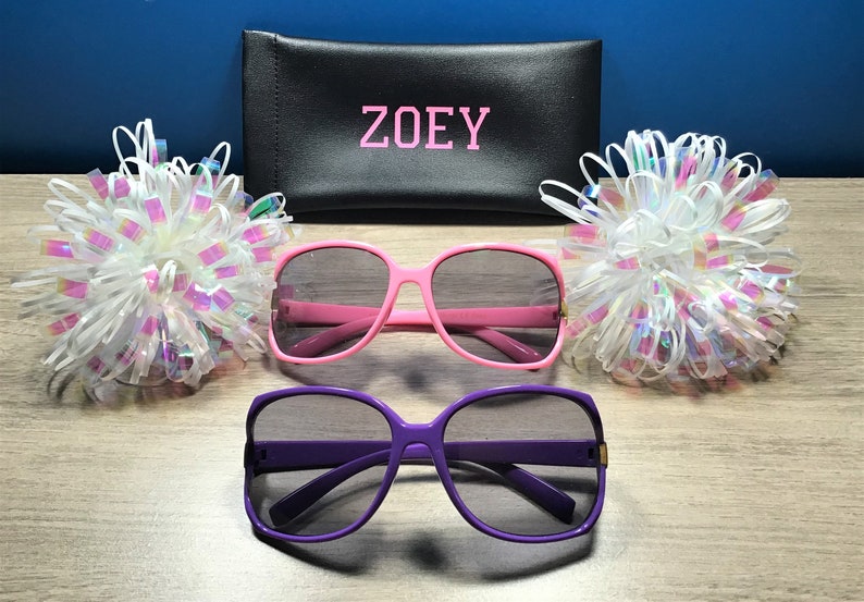 May include: Two pairs of sunglasses, one pink and one purple, with a black case that says "ZOEY" on it. The sunglasses are on a wooden surface with white and iridescent bows on either side.