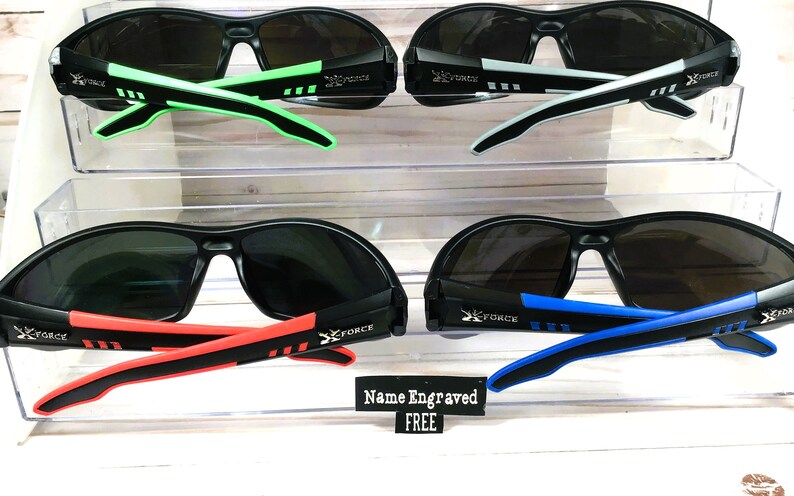 May include: Four pairs of black sunglasses with different colored accents on the arms. The sunglasses have a dark gray lens. The text "X FORCE" is printed on the arms of the sunglasses. The text "Name Engraved FREE" is printed on a white tag.