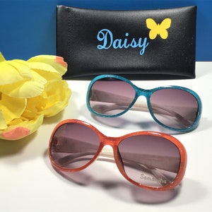 May include: Two pairs of oval-shaped sunglasses with sparkly frames. One pair is turquoise blue and the other is orange. The sunglasses have a white bridge and dark lenses. The orange pair has the name "Samantha" written on the frame.