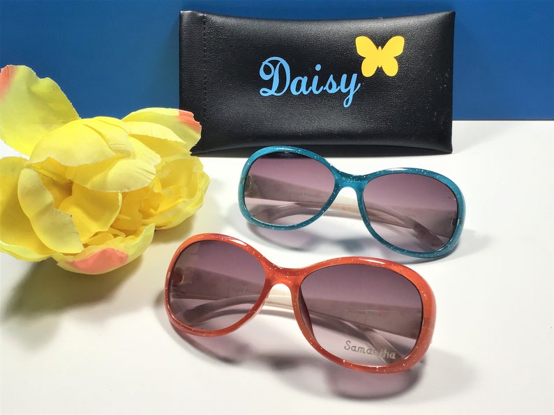 May include: Two pairs of oval-shaped sunglasses with sparkly frames. One pair is turquoise blue and the other is orange. The sunglasses have a white bridge and dark lenses. The orange pair has the name "Samantha" written on the frame.