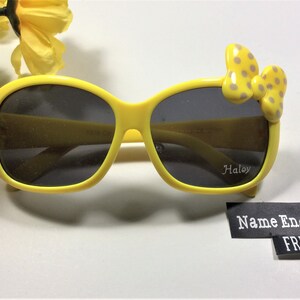 Puede incluir: Gafas de sol amarillas con un lazo de lunares en el lateral. Las gafas tienen lentes negras y el nombre "Haley" est&aacute; grabado en la lente derecha. El texto "Nombre grabado GRATIS" est&aacute; en una etiqueta negra.