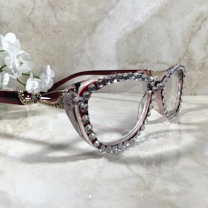 May include: Elegant eyeglasses with a burgundy frame and clear lenses. The frames are adorned with sparkling rhinestones around the lenses. The glasses are set against a marble background with white flowers.