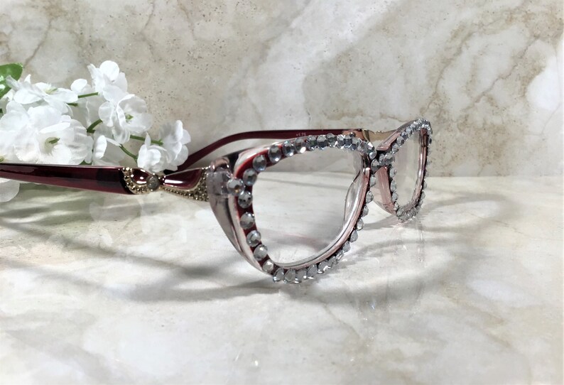 May include: Elegant eyeglasses with a burgundy frame and clear lenses. The frames are adorned with sparkling rhinestones around the lenses. The glasses are set against a marble background with white flowers.