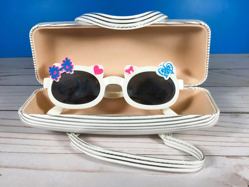 May include: White sunglasses with dark lenses, decorated with a pink heart, blue butterfly, and a blue and pink flower. The sunglasses are in a striped case with a handle. The case is white with black stripes.