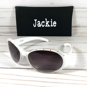 May include: White sunglasses with dark lenses and small pink accents along the top frame. Behind the sunglasses is a black rectangular case with the name Jackie printed in white text on the front. The frames feature a smooth, glossy finish.