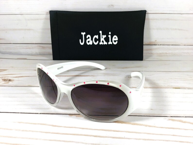 May include: White sunglasses with dark lenses and small pink accents along the top frame. Behind the sunglasses is a black rectangular case with the name Jackie printed in white text on the front. The frames feature a smooth, glossy finish.