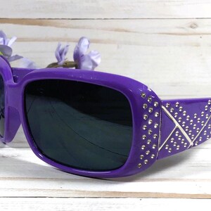 K&ouml;nnte beinhalten: Lilafarbene Sonnenbrille mit silbernen Akzenten und Strasssteinen. Die Sonnenbrille hat einen rechteckigen Rahmen und dunkle Gl&auml;ser.