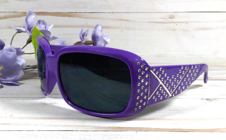 K&ouml;nnte beinhalten: Lilafarbene Sonnenbrille mit silbernen Akzenten und Strasssteinen. Die Sonnenbrille hat einen rechteckigen Rahmen und dunkle Gl&auml;ser.