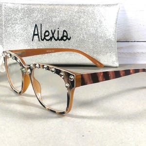 Puede incluir: Gafas con montura marr&oacute;n y lentes transparentes, adornadas con pedrer&iacute;a en el borde superior. Las patillas presentan un dise&ntilde;o a rayas marr&oacute;n y beige. Un bolso de mano plateado con el nombre "Alexia" est&aacute; en el fondo.