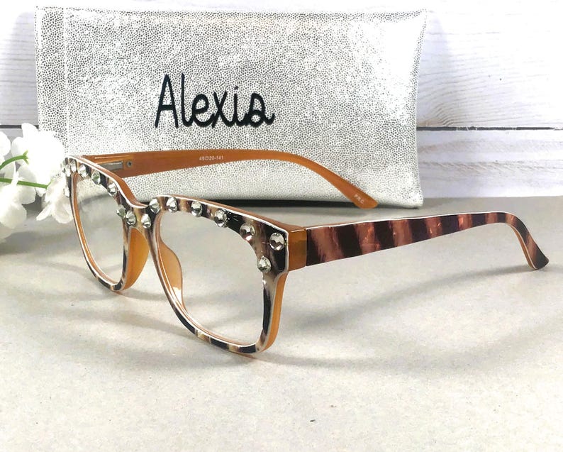 Puede incluir: Gafas con montura marr&oacute;n y lentes transparentes, adornadas con pedrer&iacute;a en el borde superior. Las patillas presentan un dise&ntilde;o a rayas marr&oacute;n y beige. Un bolso de mano plateado con el nombre "Alexia" est&aacute; en el fondo.