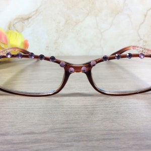 May include: Brown-framed spectacles with clear lenses, decorated with purple rhinestones along the top and on the corners. The frames feature a floral pattern on the temples. The glasses are on a light wooden surface.