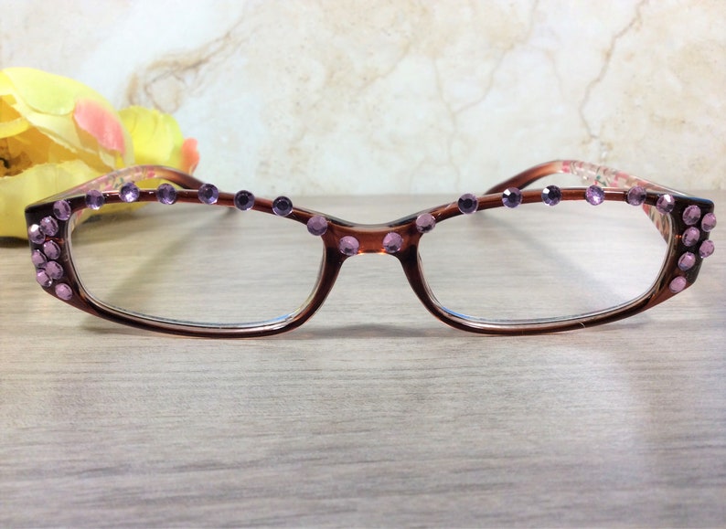 May include: Brown-framed spectacles with clear lenses, decorated with purple rhinestones along the top and on the corners. The frames feature a floral pattern on the temples. The glasses are on a light wooden surface.
