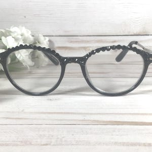 Puede incluir: Gafas negras con un dise&ntilde;o de montura redondeada y un borde festoneado texturizado a lo largo de la parte superior. Las lentes transparentes est&aacute;n colocadas en una montura de pl&aacute;stico negro brillante y las patillas son finas y del mismo color.