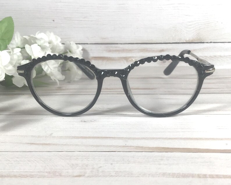 Puede incluir: Gafas negras con un dise&ntilde;o de montura redondeada y un borde festoneado texturizado a lo largo de la parte superior. Las lentes transparentes est&aacute;n colocadas en una montura de pl&aacute;stico negro brillante y las patillas son finas y del mismo color.