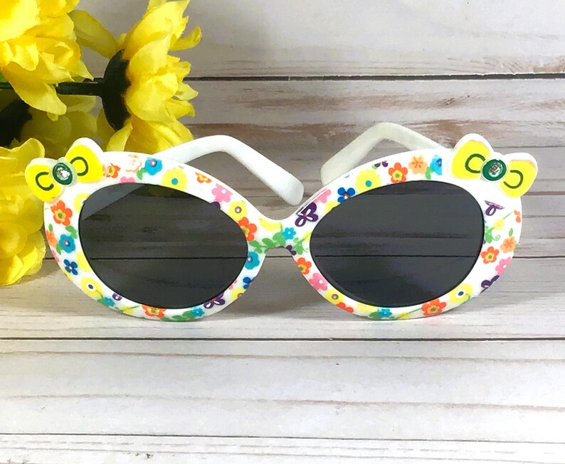 May include: White sunglasses with a floral pattern and black lenses. The sunglasses have a yellow bow on each side with the letters "CO" on them.