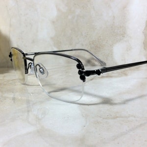 May include: A pair of eyeglasses with a black metal frame and clear lenses. The frames have a semi-rimless design with decorative black accents on the corners. The temples are thin and extend to the ear pieces.