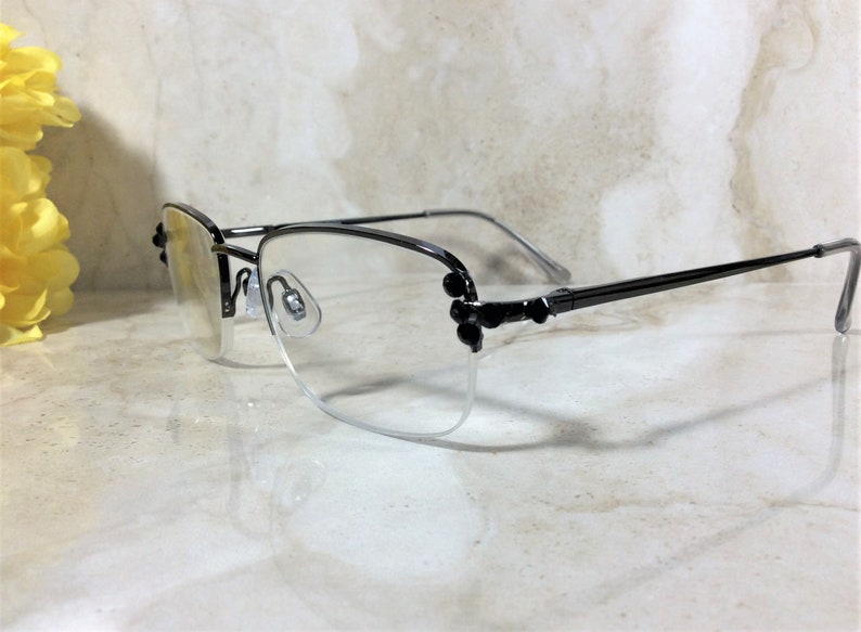May include: A pair of eyeglasses with a black metal frame and clear lenses. The frames have a semi-rimless design with decorative black accents on the corners. The temples are thin and extend to the ear pieces.