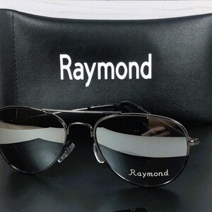May include: Aviator sunglasses with dark, reflective lenses and a thin black metal frame. The right lens features the white text 'Raymond'. Behind the sunglasses sits a black rectangular protective case with the white text 'Raymond' printed on it.