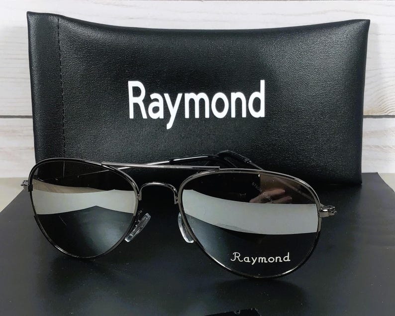 May include: Aviator sunglasses with dark, reflective lenses and a thin black metal frame. The right lens features the white text 'Raymond'. Behind the sunglasses sits a black rectangular protective case with the white text 'Raymond' printed on it.