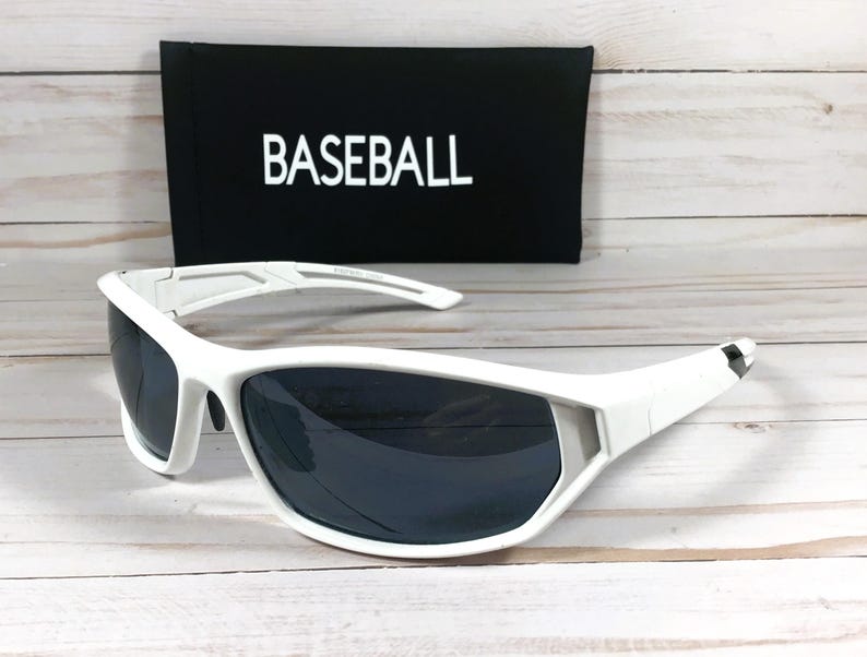 May include: White sports sunglasses with dark lenses are displayed in front of a black case. The case has the word "BASEBALL" printed in white. The sunglasses have a sporty design.