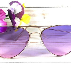 May include: Gold-framed aviator sunglasses with gradient purple lenses. The sunglasses are set against a backdrop of white wood and colourful flowers, creating a stylish and eye-catching accessory.