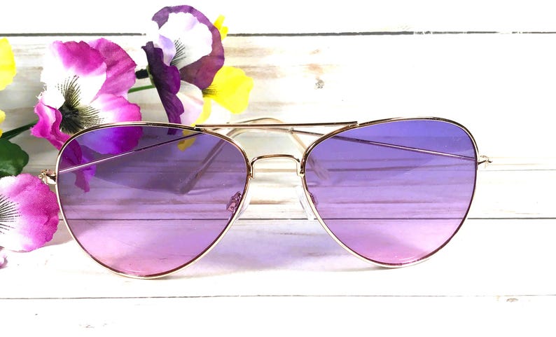 May include: Gold-framed aviator sunglasses with gradient purple lenses. The sunglasses are set against a backdrop of white wood and colourful flowers, creating a stylish and eye-catching accessory.