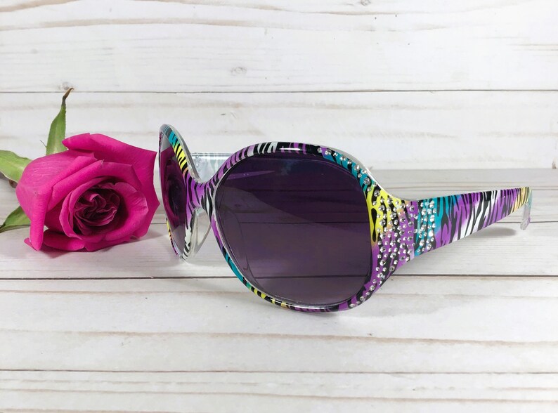 May include: Purple, yellow, and blue zebra print sunglasses with rhinestones on the frame. The sunglasses have dark lenses.