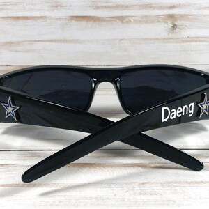 May include: Black sunglasses with a rectangular frame. The temples feature a silver star and the word "Daeng" in white. The lenses are dark, and the sunglasses are set against a light wood background.