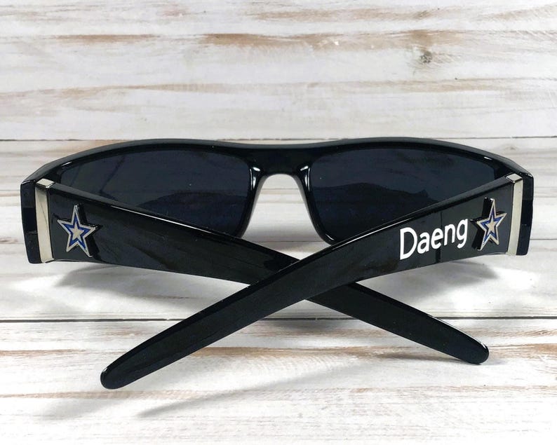 May include: Black sunglasses with a rectangular frame. The temples feature a silver star and the word "Daeng" in white. The lenses are dark, and the sunglasses are set against a light wood background.