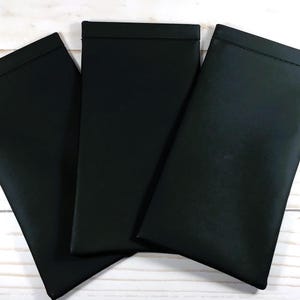 May include: Three black fabric pouches with a stitched edge. The pouches are lying flat on a white wooden surface.