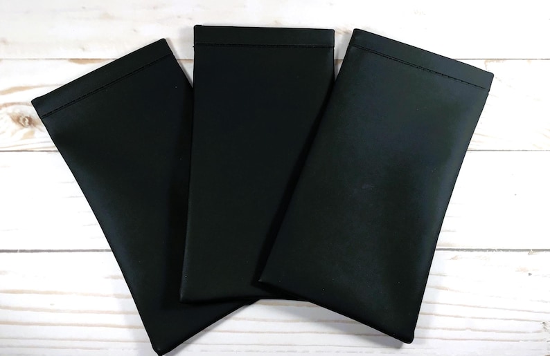 May include: Three black fabric pouches with a stitched edge. The pouches are lying flat on a white wooden surface.