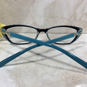 May include: Black and teal eyeglasses with a cat-eye frame. The glasses have teal temples and decorative accents at the hinges. The lenses are clear. The glasses are set against a neutral background.