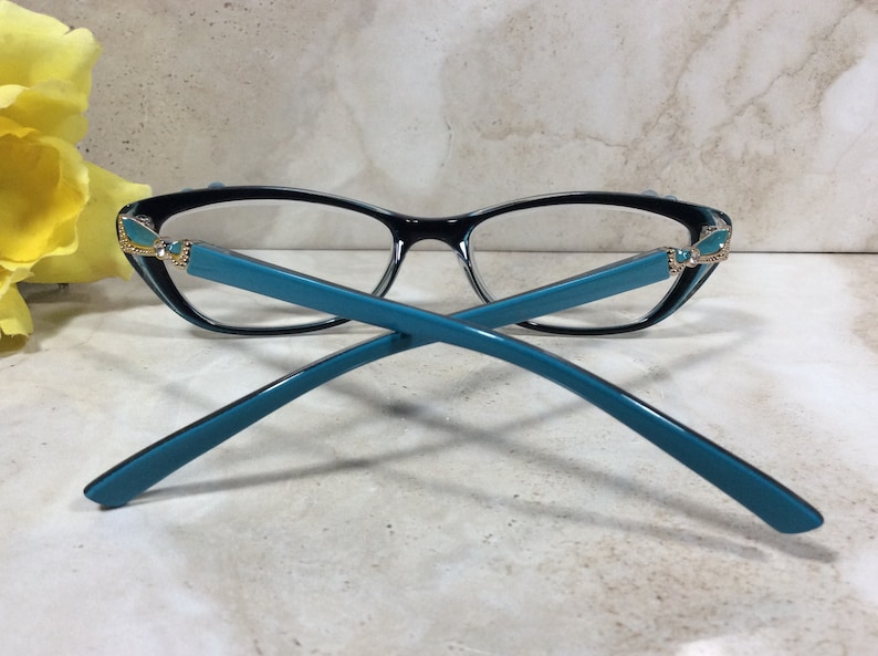 May include: Black and teal eyeglasses with a cat-eye frame. The glasses have teal temples and decorative accents at the hinges. The lenses are clear. The glasses are set against a neutral background.
