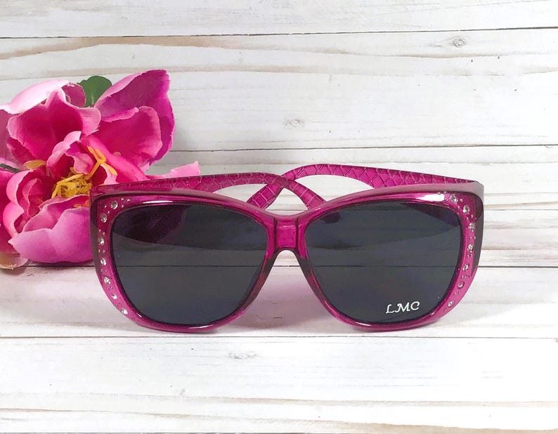 May include: Hot pink sunglasses with dark lenses and a textured frame. The sunglasses are embellished with small rhinestones along the top edge of the frame. The brand name "LMC" is printed on the right lens.