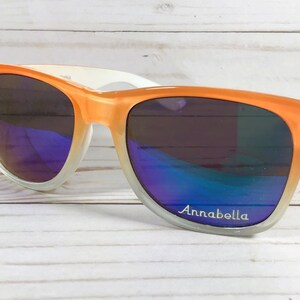 Puede incluir: Gafas de sol de color naranja y blanco degradado con una inscripci&oacute;n plateada que dice "Annabella".
