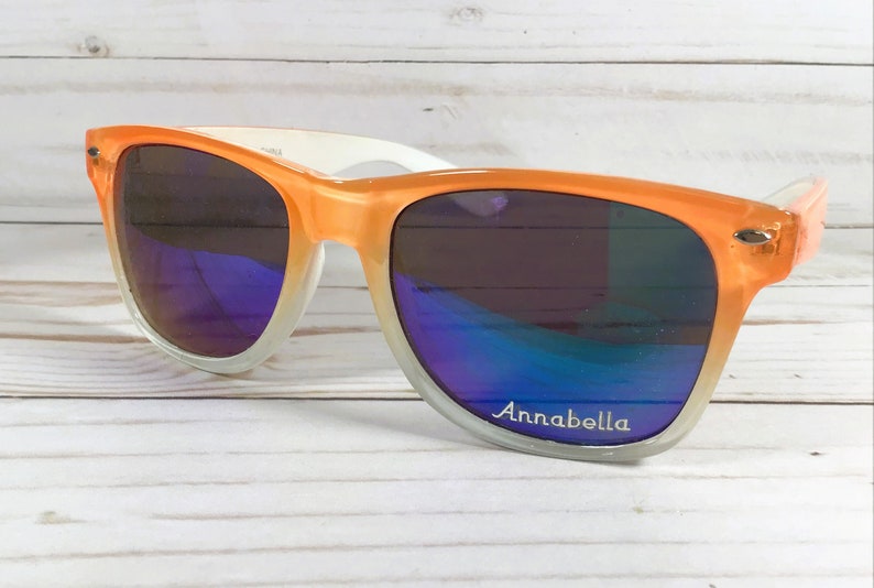 Puede incluir: Gafas de sol de color naranja y blanco degradado con una inscripci&oacute;n plateada que dice "Annabella".