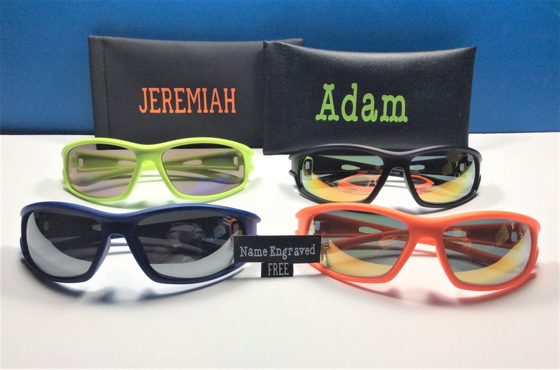 Puede incluir: Cuatro pares de gafas de sol con diferentes colores de montura y lentes. Las gafas de sol est&aacute;n dispuestas en dos filas de dos. La fila superior presenta un par de gafas de sol verdes con una funda negra etiquetada como "JEREMIAH" y un par de gafas de sol negras con una funda negra etiquetada como "Adam". La fila inferior presenta un par de gafas de sol azules y un par de gafas de sol naranjas. Un cartel en el medio de la imagen dice "Nombre grabado GRATIS".