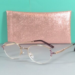May include: Gold-rimmed spectacles with oval lenses and purple accents. The glasses are set against a rose gold glitter clutch. The frames have a delicate design, and the lenses are clear.